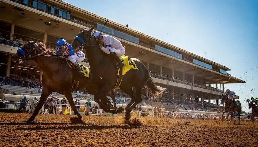 Jockeys on horseback race down a dirt track in front of a large, sunlit grandstand filled.