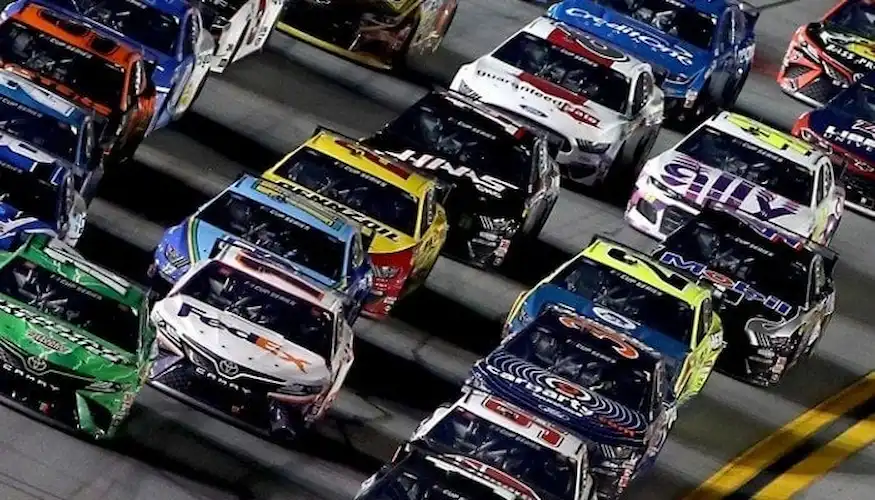 A tight pack of NASCAR race cars speeds down the track under the lights during a high-intensity race.