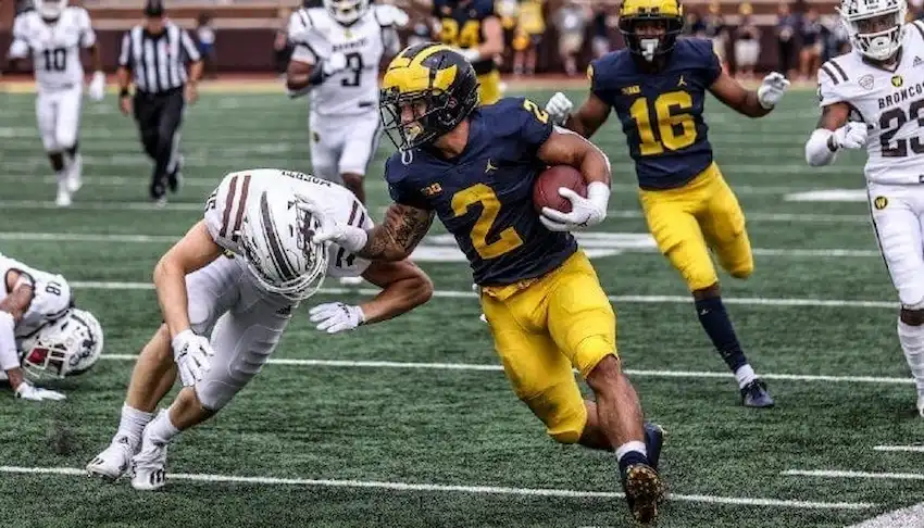 Gambling News 11 A college football player runs with the ball while stiff-arming a defender during a game.