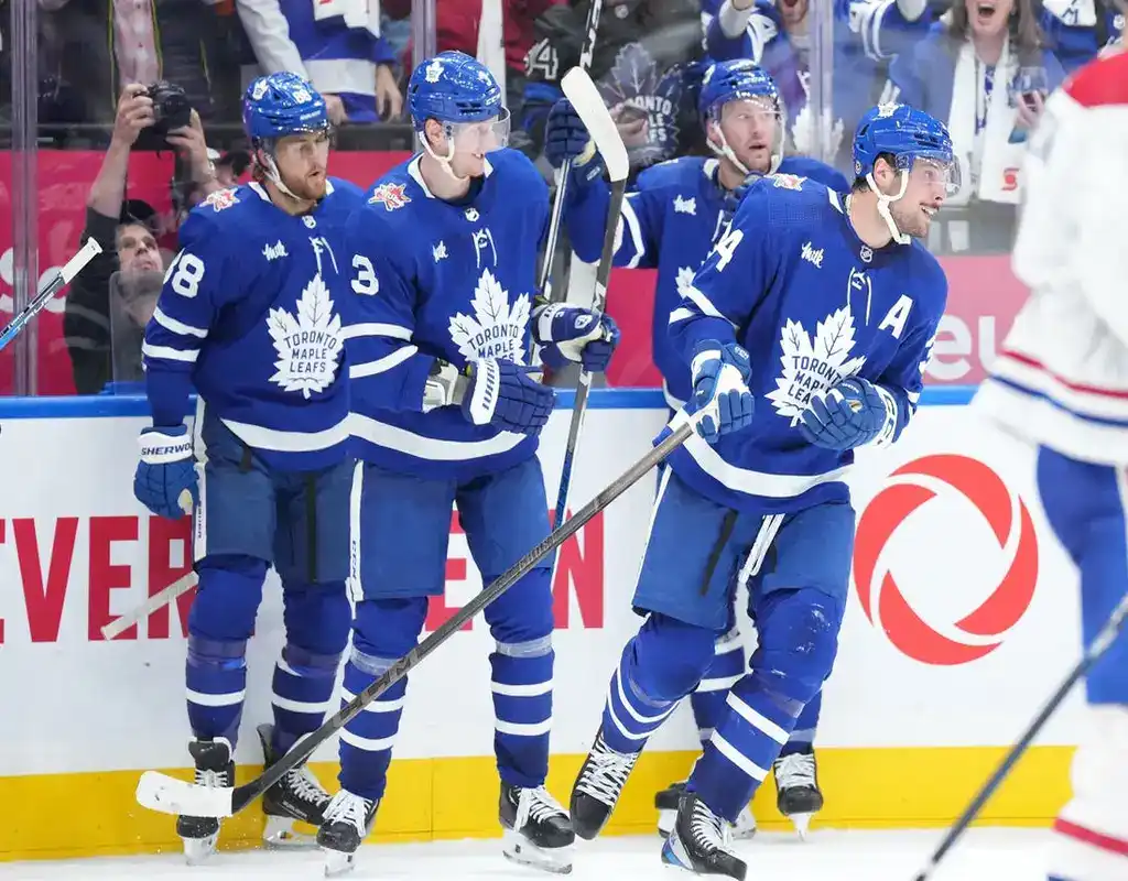 Toronto Maple Leafs players clustered in celebration along the boards with a Minnesota Wild player visible on the ice