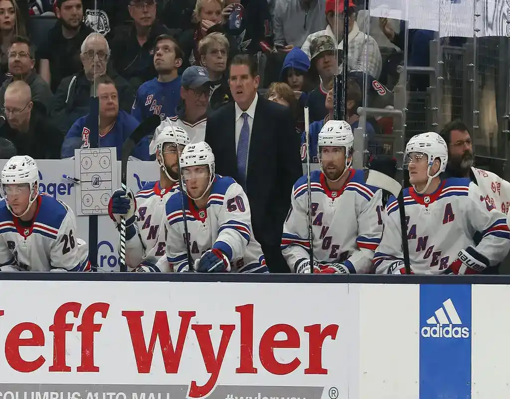 New York Rangers players and coaching staff watch the game intently from the bench during a match against the Carolina Hurricanes, with fans visible behind the glass.