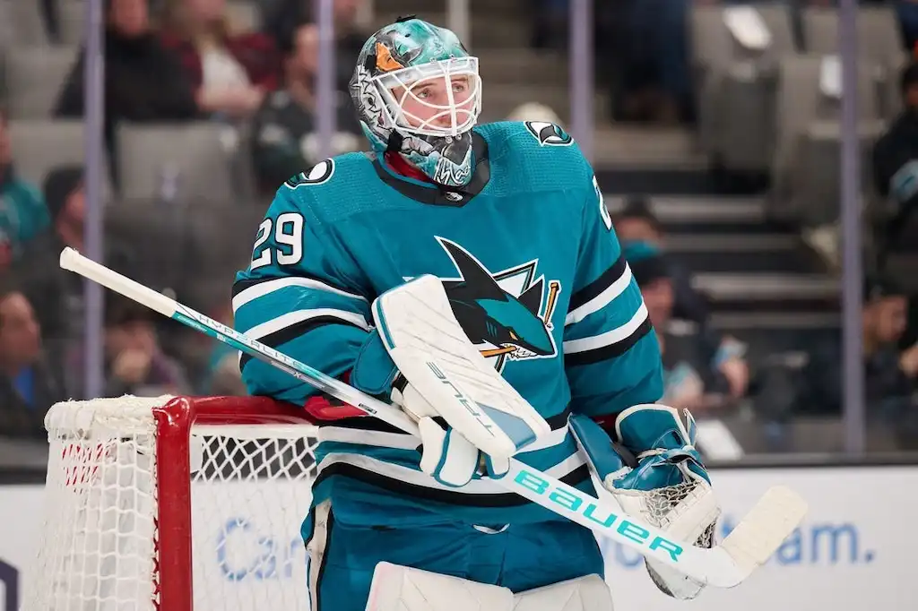NHL 4 A San Jose Sharks goalie in teal gear and a decorated helmet stands in front of the net during a game.