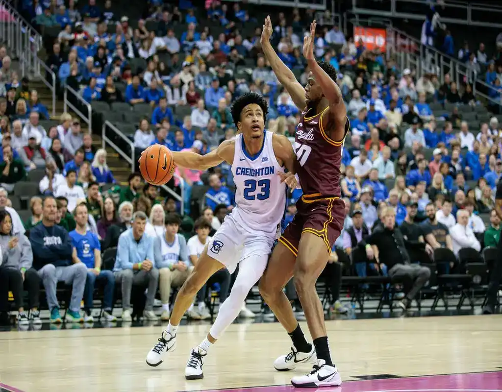 Creighton Bluejays #23 protecting the ball as Colorado State Rams defender #10 raises his arms in a crowded arena