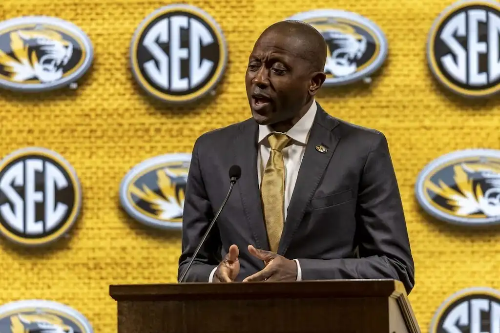 Missouri Coach speaks at a podium, possibly during a press conference or official announcement.