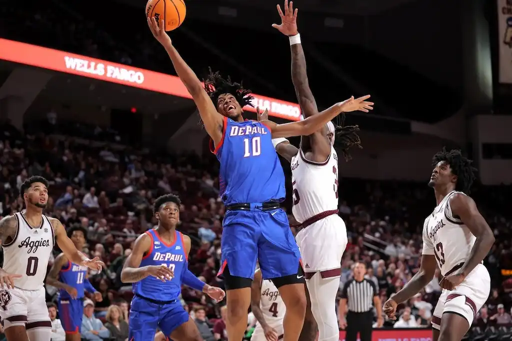 A DePaul player leaps toward the basket for a layup while being closely contested by a Texas A&M defender.