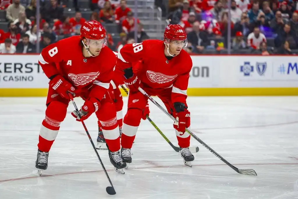Two Detroit Red Wings players skate side by side during a game, focused on the puck as they move up the ice.