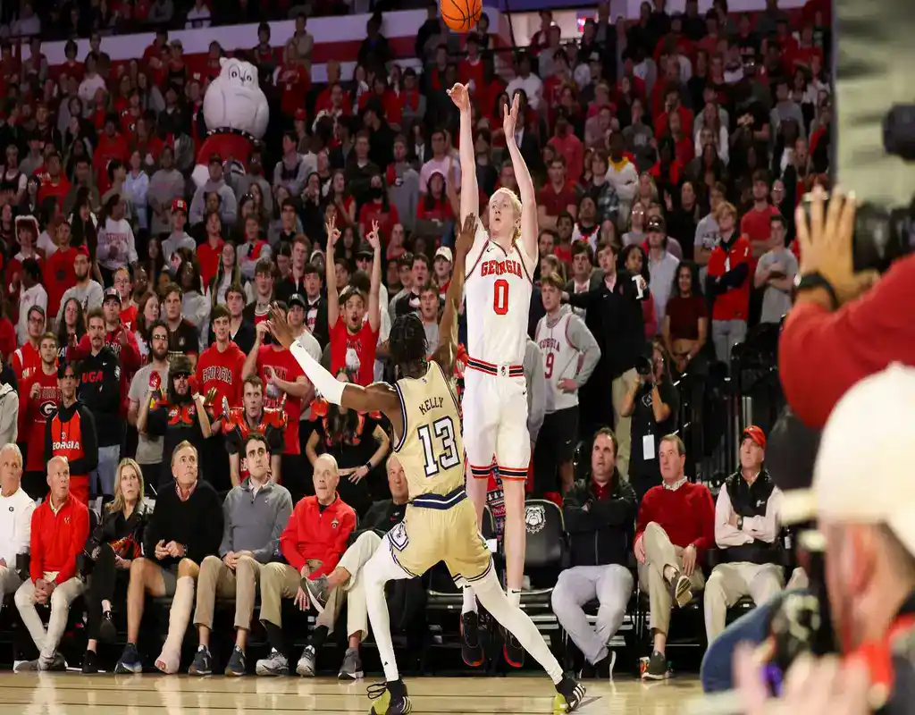 Georgia Bulldogs player #0 leaping for a jump shot over Alabama A&M defender #13 in front of the crowd