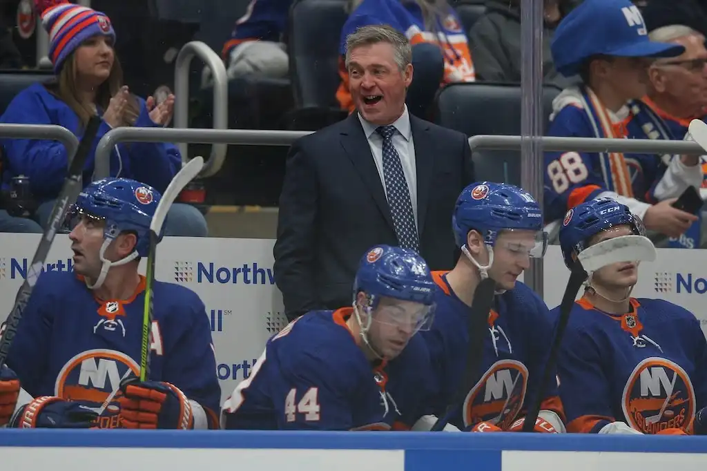 New York Islanders coach stands behind the bench, smiling.