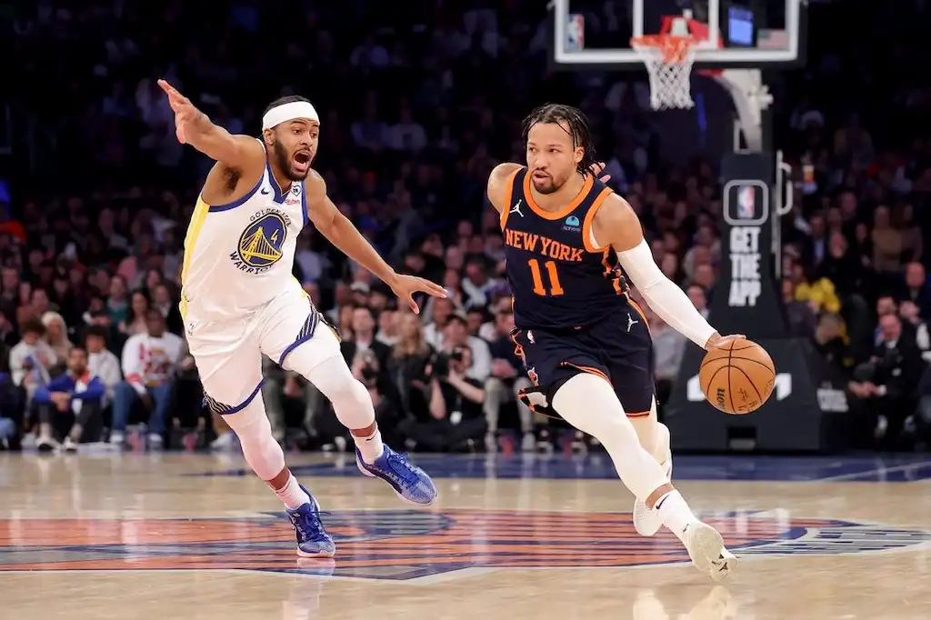 NY Knicks player dribbles the basketball up the court while being closely defended by a GS Warriors player.