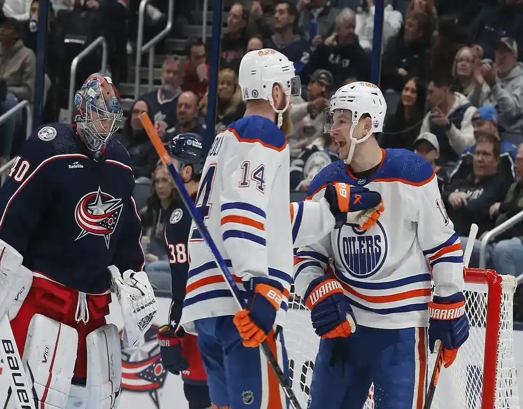 Edmonton Oilers players #14 and #19 celebrating near the goal crease as Columbus Blue Jackets goalie #40 stands ready in an NHL arena