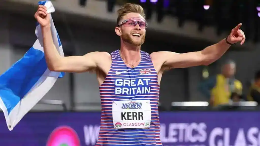 Athlete in a striped Great Britain outfit celebrates triumphantly, waving a Scottish flag and pointing confidently at the crowd.