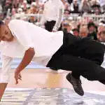A breakdancer in a white shirt and black pants performs a dynamic move on stage, with an engaged audience in the background.