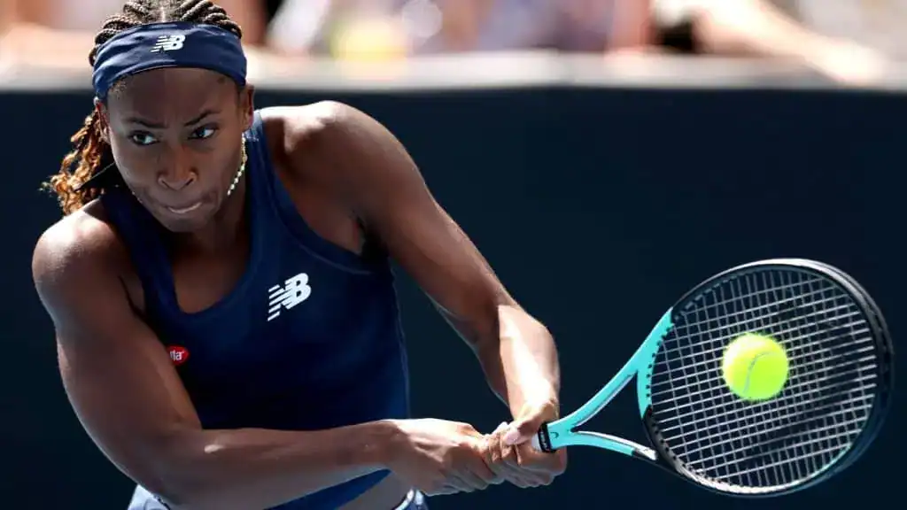 A tennis player in a dark sleeveless shirt prepares to hit a yellow ball with a blue and black racket during a match.