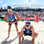 Two beach volleyball players celebrate energetically on the sand, with a cheering crowd and flags in the background under a bright sky.