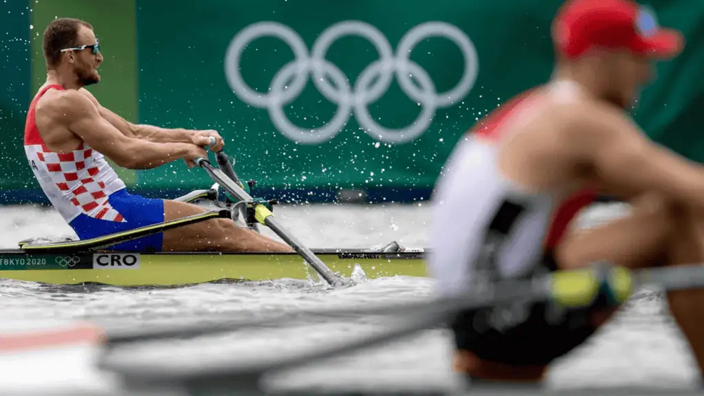 A Croatian rower races in a single scull on the water, Olympic rings in the background, with droplets splashing around.