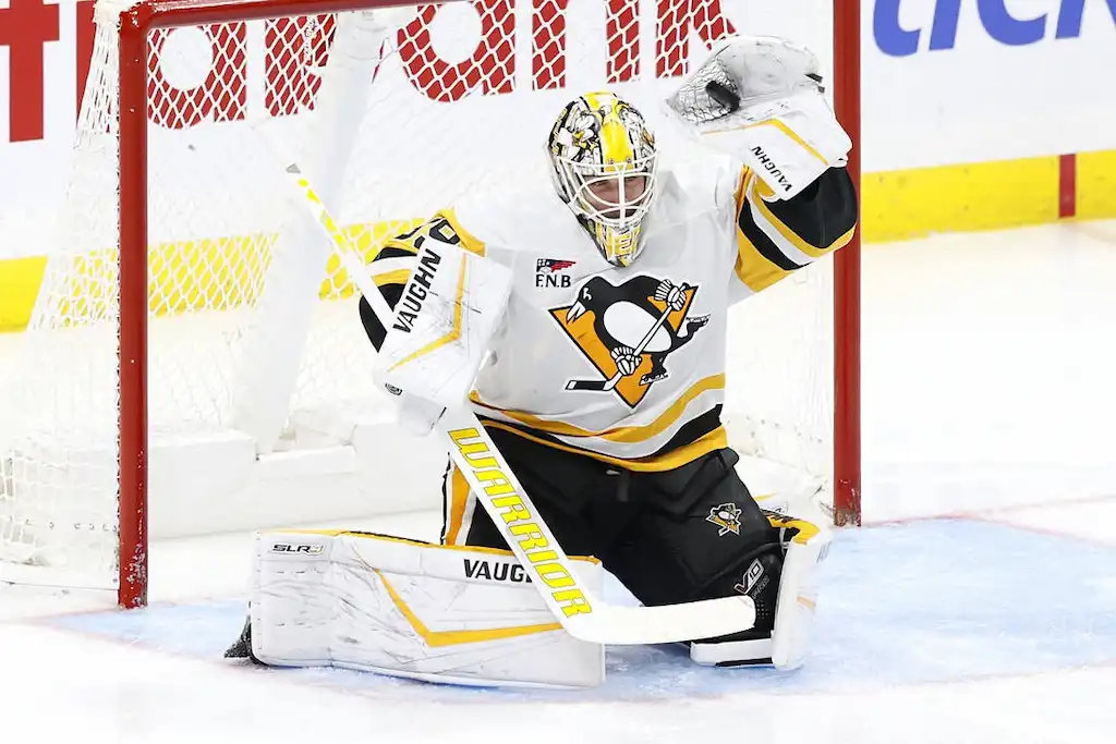 A Pittsburgh Penguins goalie in full gear makes a save while kneeling in front of the net.