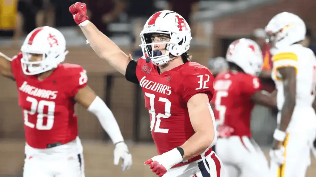 A football player in a red Jaguars uniform celebrates a key play, raising his arm with excitement as teammates react in the background.