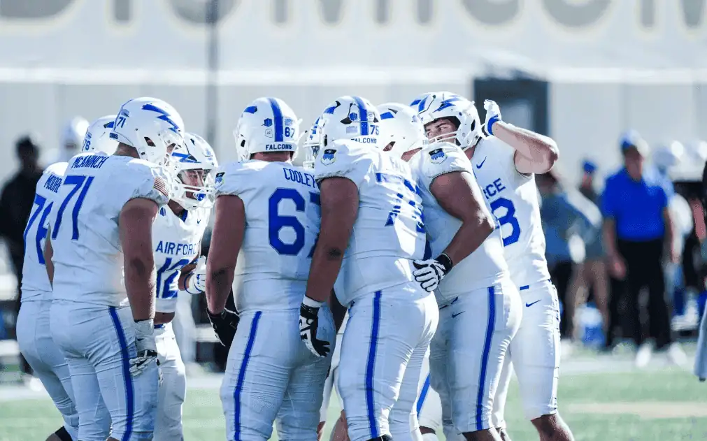 A group of football players in white uniforms with blue accents huddle on the field, discussing strategy during a game.