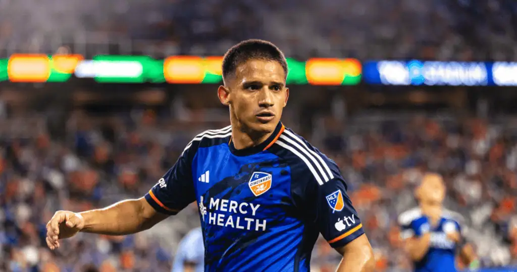 A soccer player wearing a blue and orange jersey celebrates in front of an energized crowd during a night game.