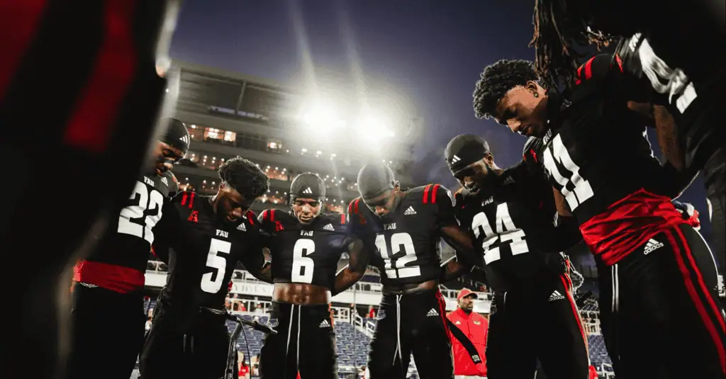 NCAAF 6 A group of football players in black and red uniforms huddle together under bright stadium lights, showing unity and focus.