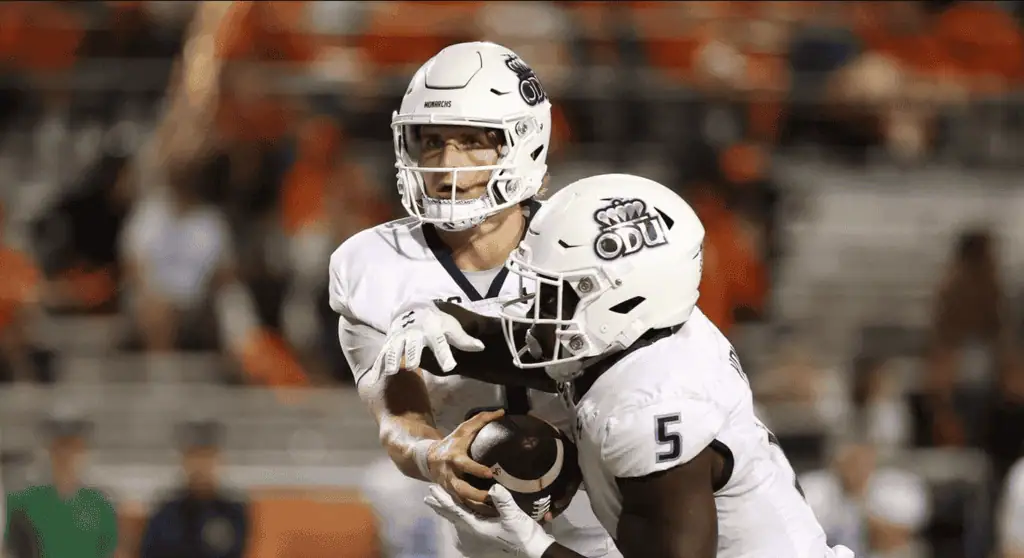 A football player in a white jersey receives a handoff during a game, with an enthusiastic crowd in the background.