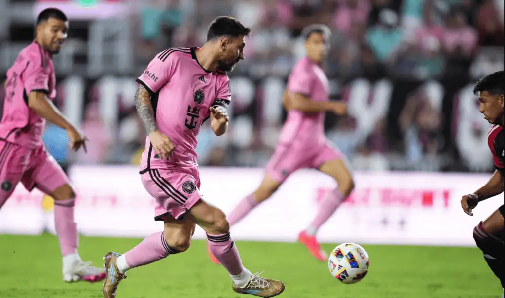 A soccer player in a pink jersey skillfully dribbles the ball during an intense game, with teammates and spectators in the background.