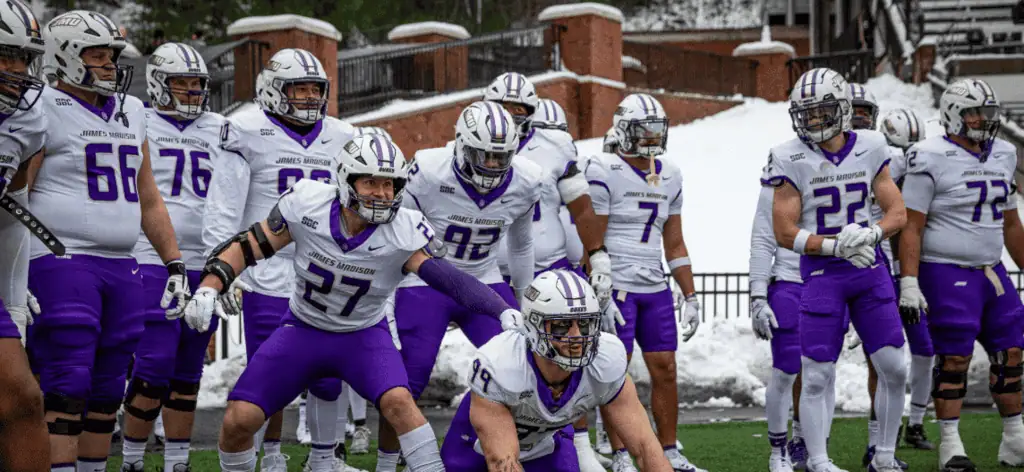 A group of football players in white and purple uniforms prepares for a play on a snowy field, showcasing teamwork and focus.