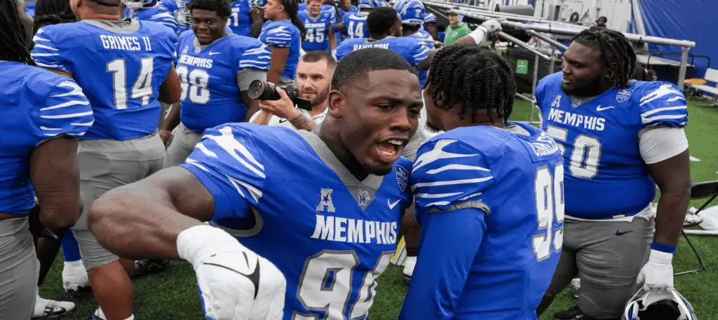 Football players in blue Memphis jerseys celebrate and strategize on the sidelines, showcasing teamwork and excitement.