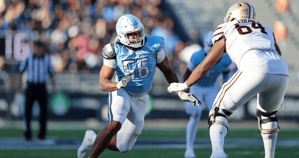 A football player in a light blue uniform tackles an opponent in white during a game, with spectators in the blurred background.