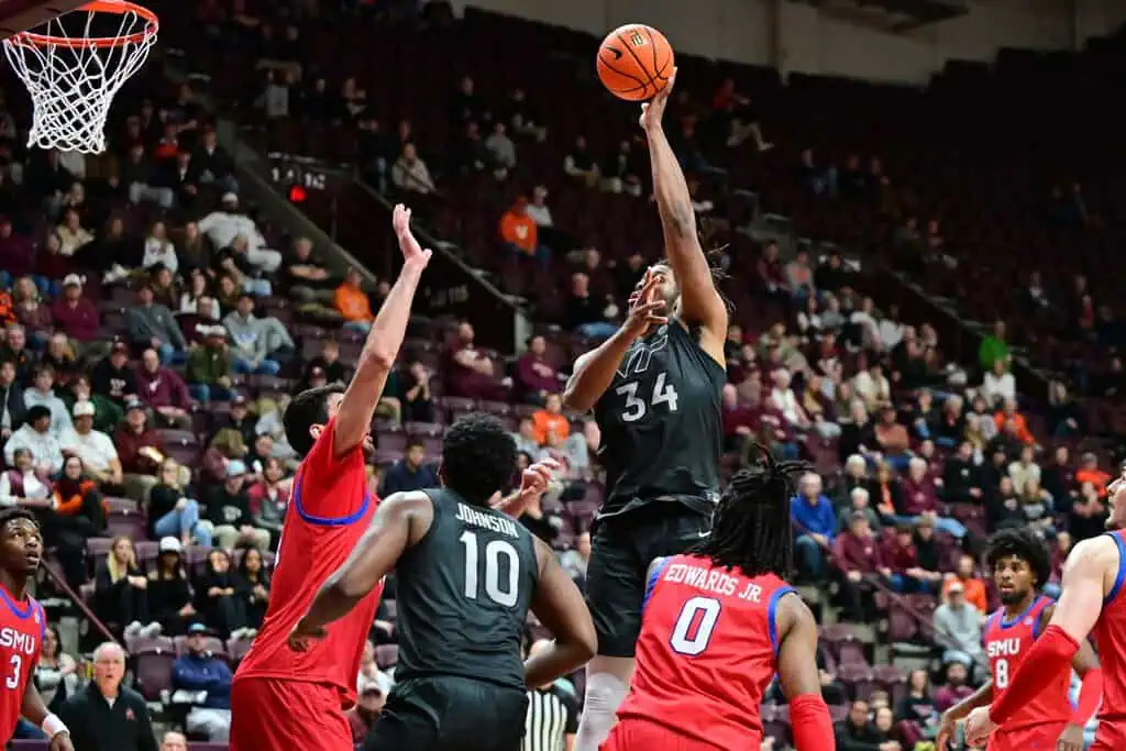 A basketball player in a black uniform leaps to shoot while two opponents in red attempt to block the shot during a game.