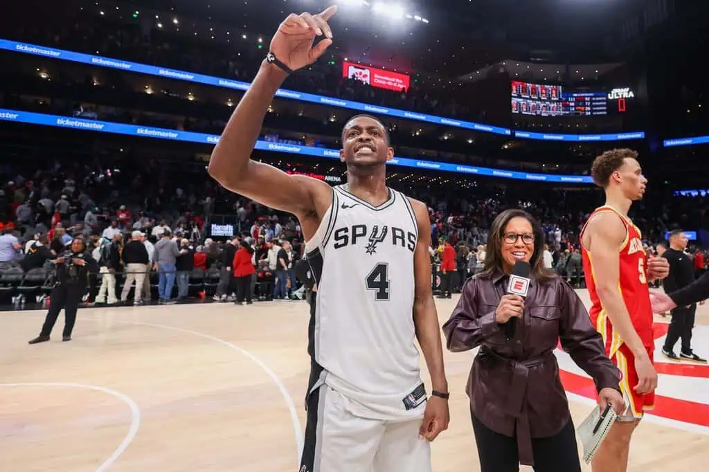 A San Antonio Spurs player gestures to the crowd while an ESPN reporter conducts an interview post-game, with a cheering audience behind.