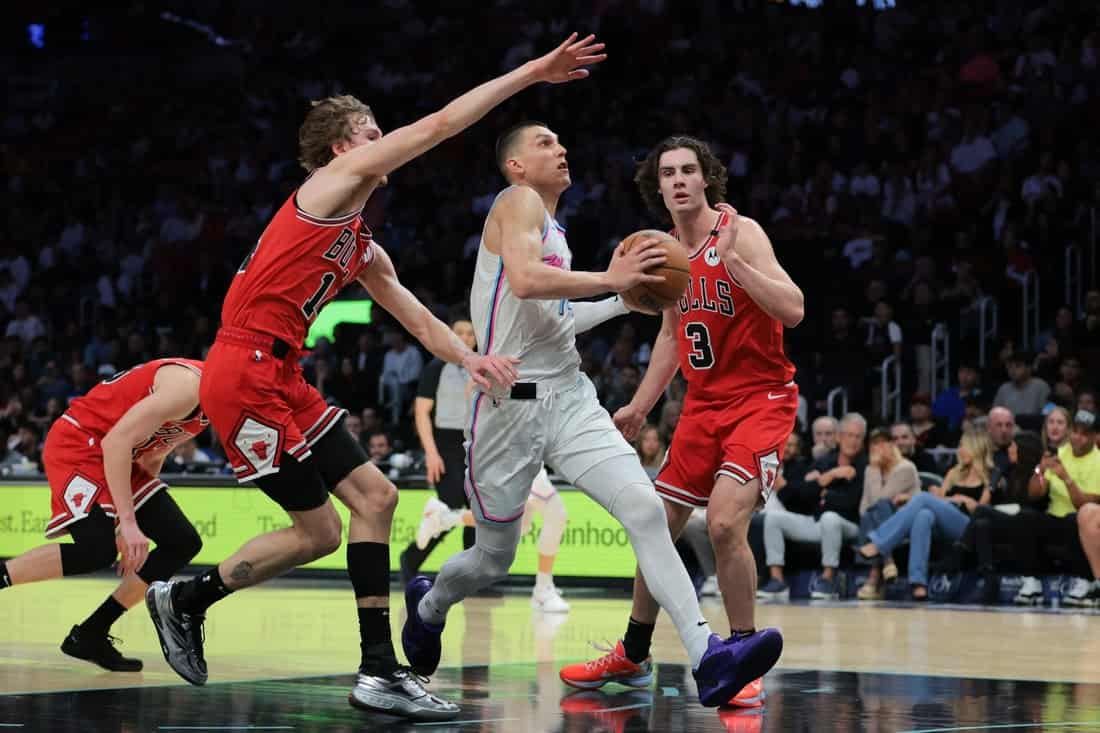 9 A player in a white jersey dribbles past two defenders in red jerseys during an intense basketball game, with spectators in the background.