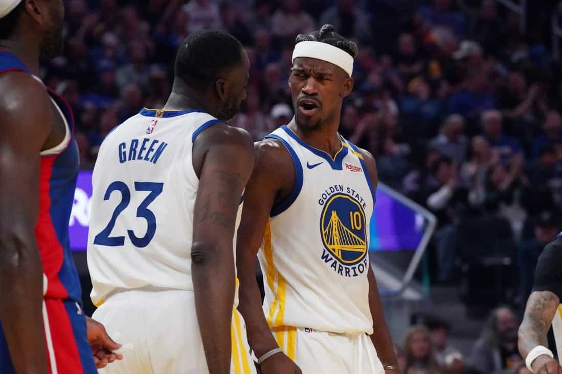 Two basketball players in Golden State Warriors uniforms are on the court. One looks animated and expressive, while the other faces him, appearing calm.
