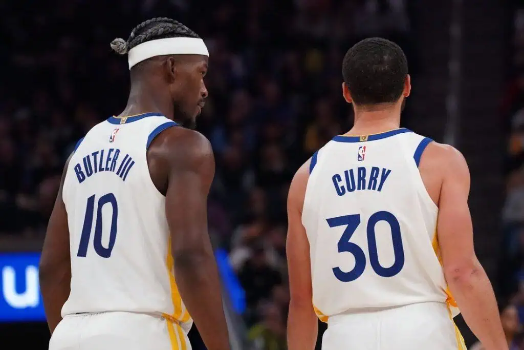 Two basketball players stand back-to-back on the court, wearing golden state warrior jerseys with the names "Butler III" and "Curry."