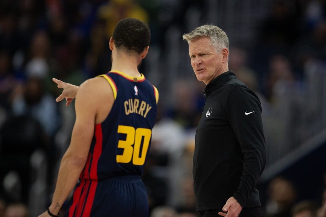 10 NBA player Stephen Curry, wearing a Golden State Warriors jersey, gestures while engaging in conversation with a coach during a game.