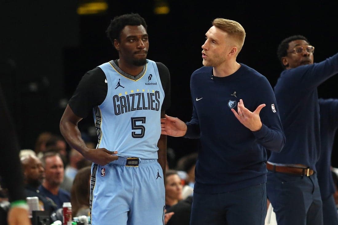 A Memphis Grizzlies player, wearing a light blue jersey, interacts with a coach during a basketball game on the sidelines.