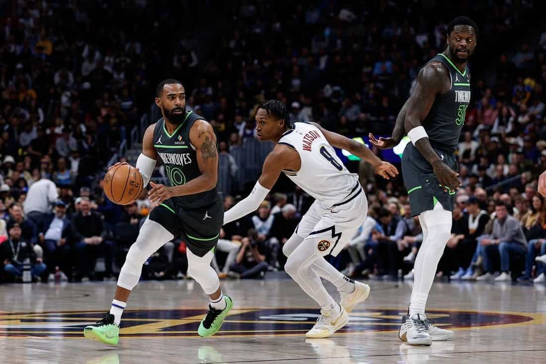Two basketball players from opposing teams in action on the court. One dribbles the ball while the other defends. The crowd watches intently.