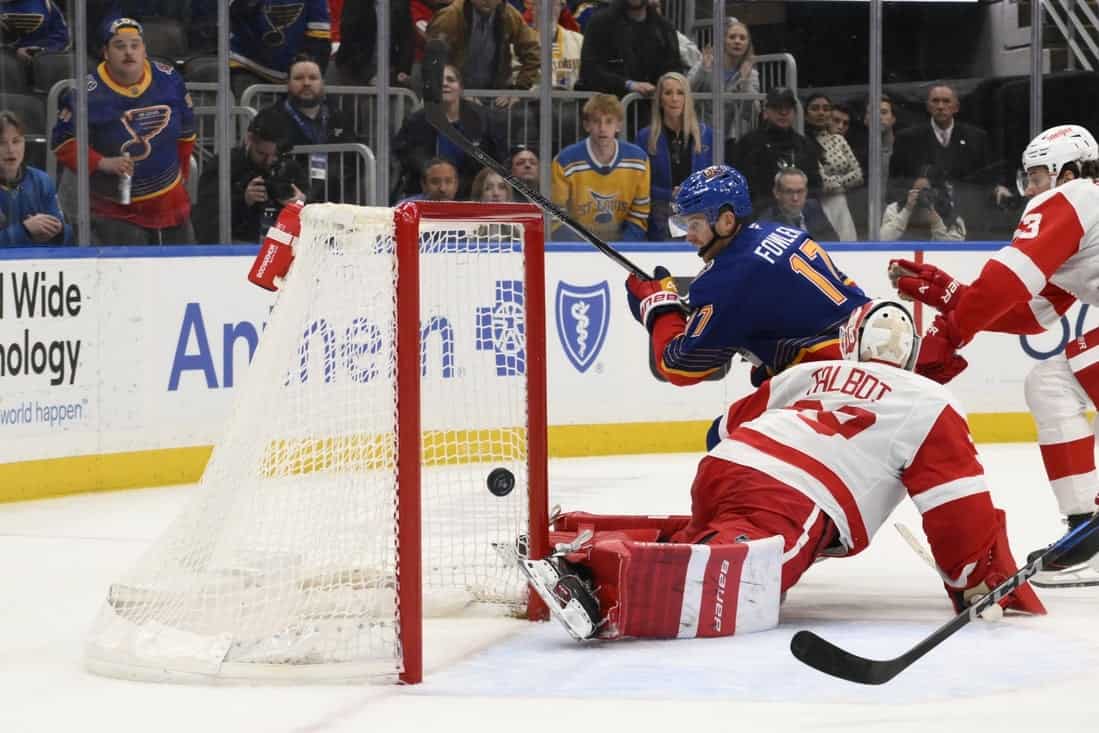 A hockey player in a blue jersey shoots the puck as the opposing goalie in red dives to block it, amidst an excited crowd.