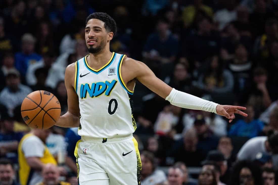 A basketball player in a white and blue "INDY" jersey dribbles a ball, poised to make a play on the court surrounded by fans.