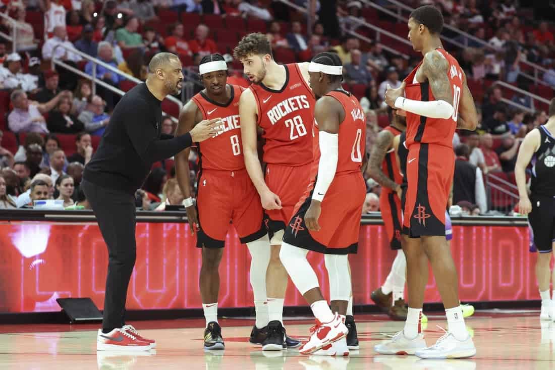 A coach engages with four Houston Rockets players, all in red uniforms, during a basketball game with fans in the background.