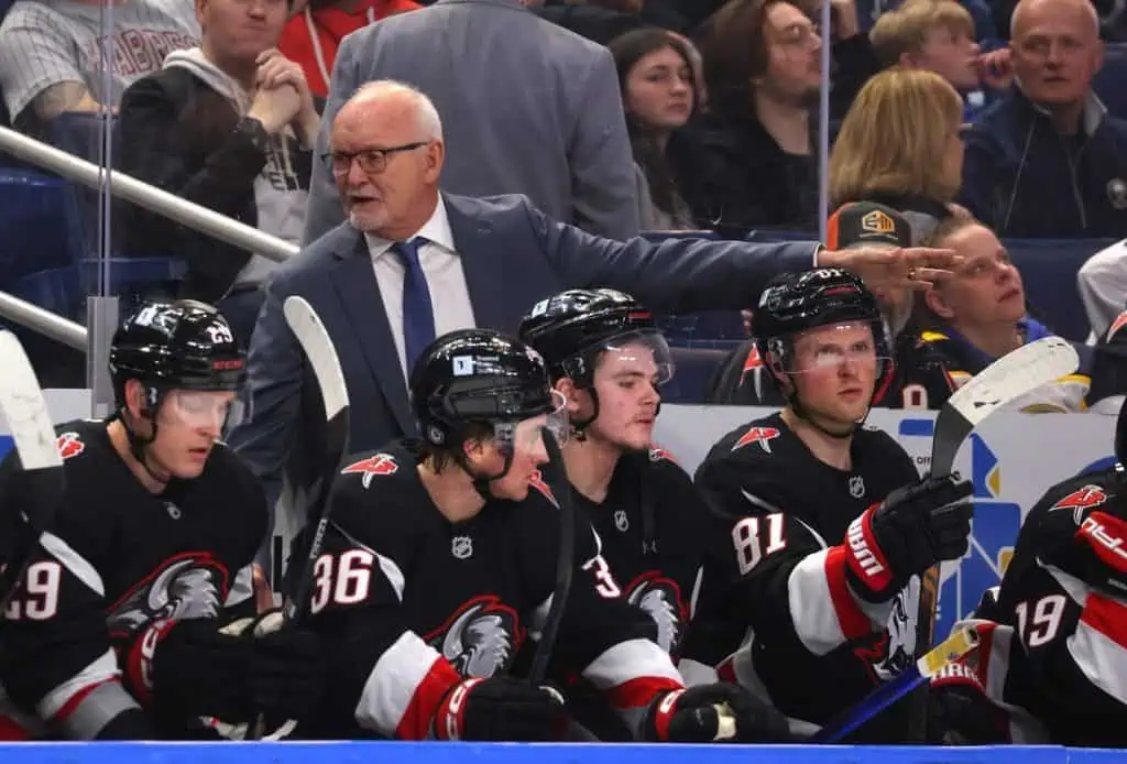 A hockey coach gestures from the bench while players in black jerseys focus on the game during a lively match in a stadium.