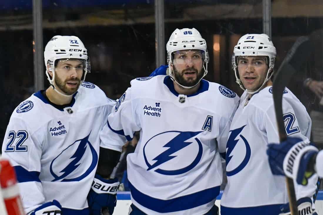 Three hockey players in white jerseys with a lightning bolt logo celebrate on the ice, showcasing teamwork and camaraderie.