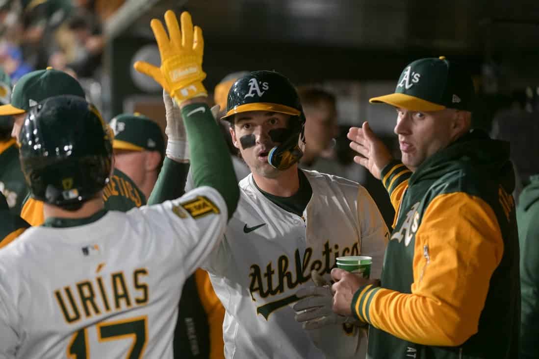 Baseball players celebrate in the dugout, wearing Oakland Athletics uniforms, sharing excitement after a play.