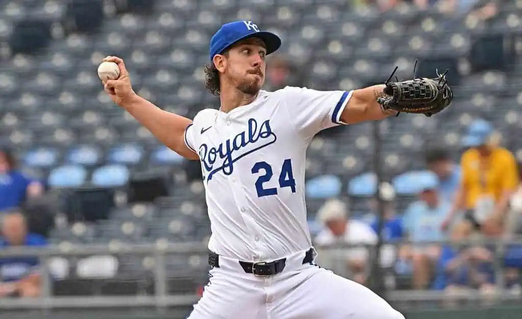 A pitcher in a white Kansas City Royals uniform winds up to throw a baseball during a game, with fans visible in the blurred background.