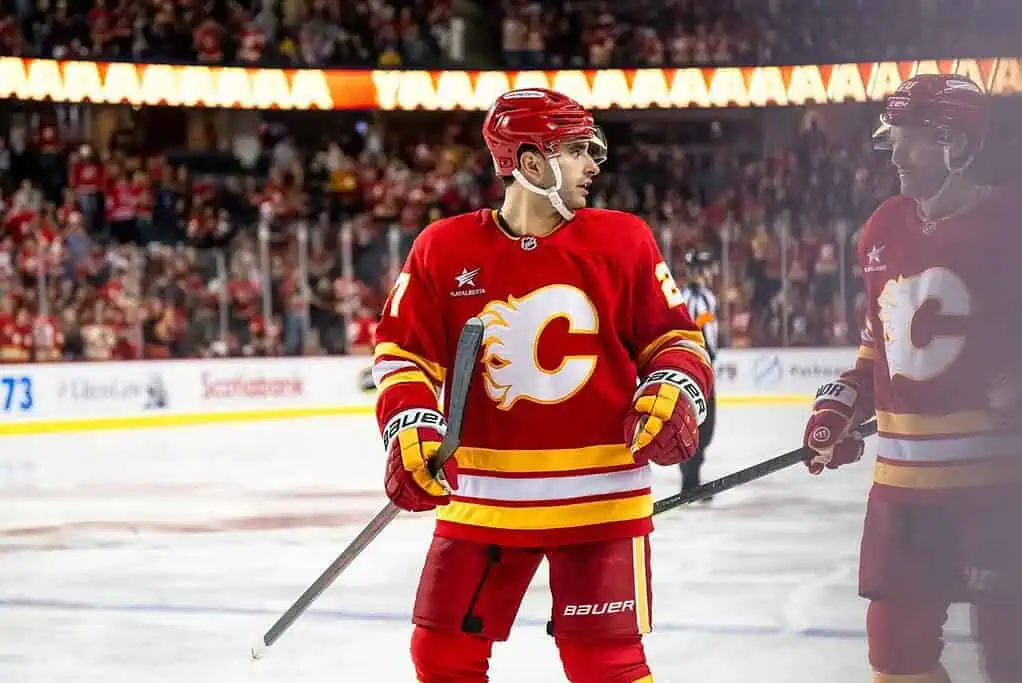 A Calgary Flames player in a red jersey stands on the ice, with fans cheering in the background during an intense hockey game.