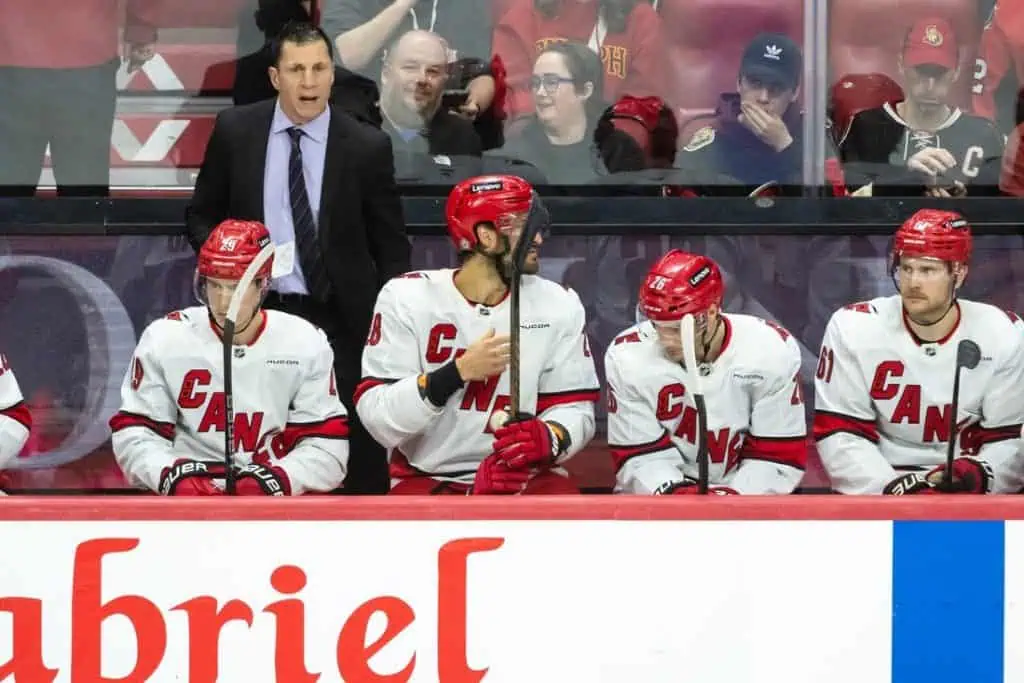 A hockey team sits on the bench wearing red and white jerseys while a coach stands nearby, focused on the game.