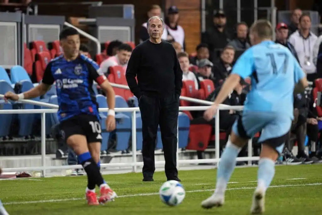 A soccer match in progress with players in blue and black uniforms, a coach overseeing from the sidelines, and spectators in the stands.