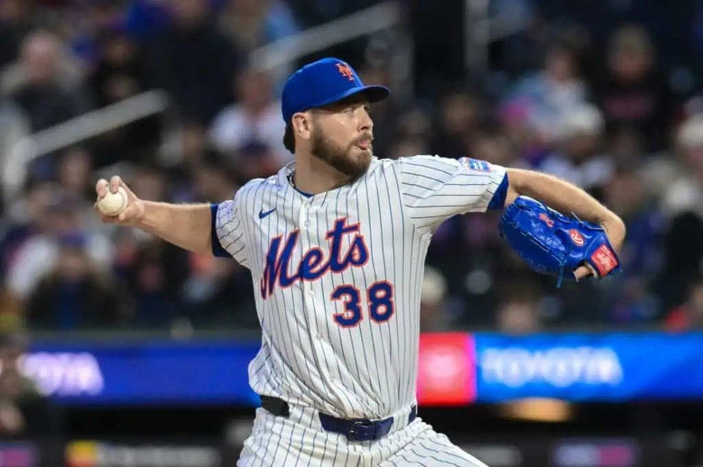 A New York Mets pitcher in a white striped uniform winds up to throw a baseball, with fans visible