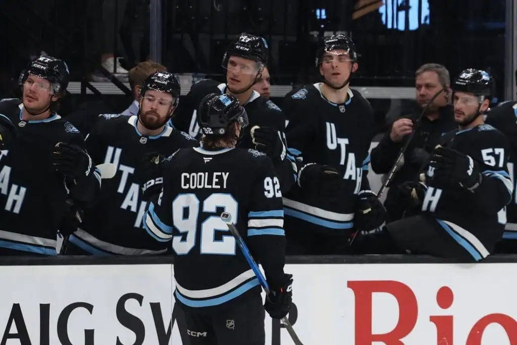 A hockey player wearing a black jersey with "COOLEY 92" stands on the ice, while teammates celebrate and cheer from the bench.