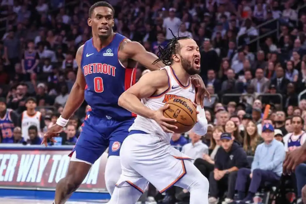 A basketball player in a white Knicks jersey dribbles the ball while being closely guarded by an opponent in a blue Pistons jersey.
